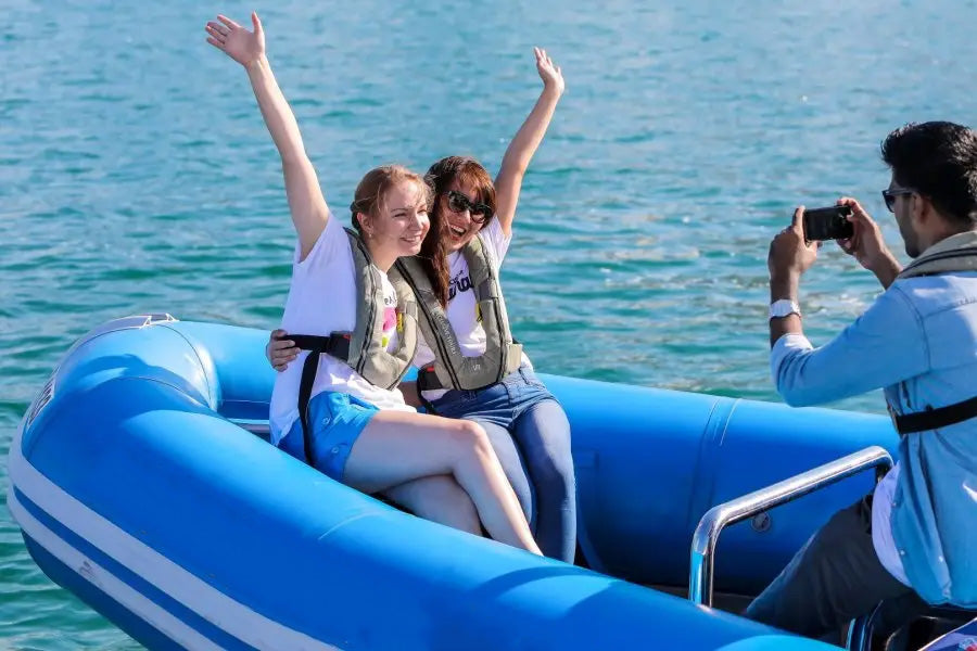 Two people on an inflatable boat with a third person taking a photo, all against a blue water background.
