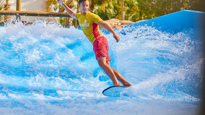 Person surfing on a blue wave simulator with a yellow shirt and red shorts.