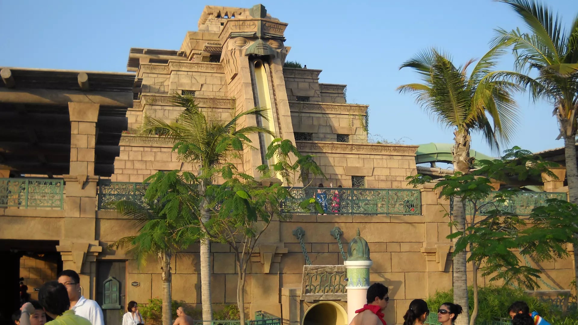 Architectural structure with a water slide, surrounded by palm trees and people.