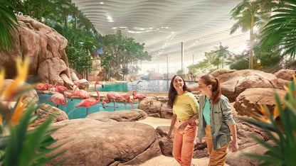 Two women walking through a tropical-themed indoor attraction with flamingo exhibits.