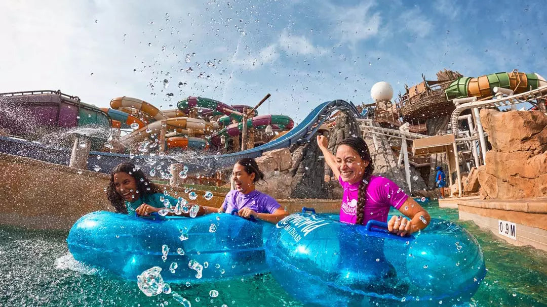 Three people on inflatable rafts in a water park with slides and attractions in the background.
